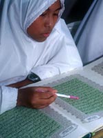 A girl wearing a white gown and Muslim headscarf and digital watch reads the Qur'an