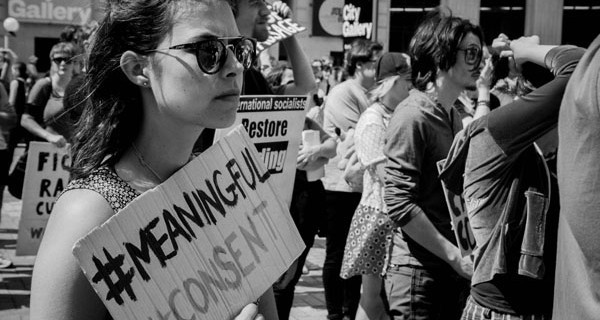 A young woman holds a sign reading "#meaningful #consent" in a protest