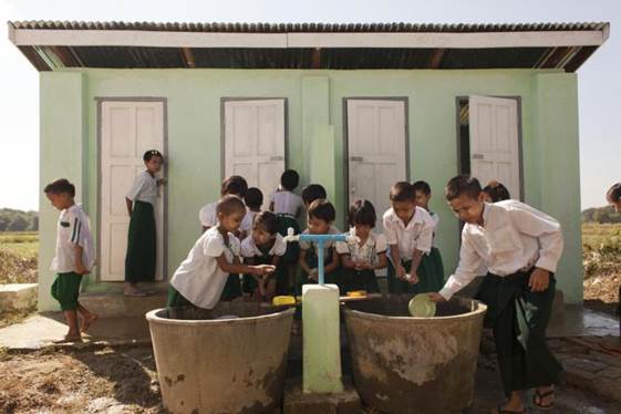Children in Myanmar wash their hands with soap at a hand-washing station, while other students behind them wait their turn to use latrines.